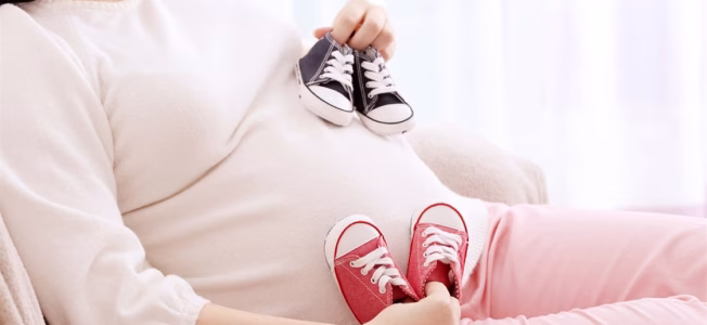صورة لسيدة في شهور الحمل الأولى تحمل أحذية لأطفالها المستقبليين بعد أن قرأت عن تغذية التوائم اثناء الحمل